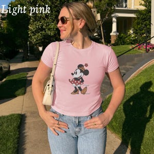 May include: Light pink t-shirt featuring a Minnie Mouse graphic. The graphic shows Minnie Mouse in a red polka dot dress and brown shoes. The shirt has short sleeves and a crew neck. The words "Light pink" are in the top left corner.