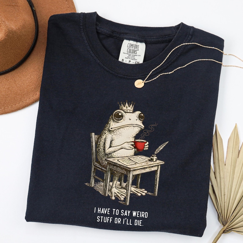 K&ouml;nnte beinhalten: Marineblaues T-Shirt mit einer skurrilen Illustration einer Froschk&ouml;nigin, die an einem Schreibtisch sitzt und eine rote Tasse h&auml;lt. Der Text "I HAVE TO SAY WEIRD STUFF OR I'LL DIE" ist darunter gedruckt. Eine goldene Halskette liegt auf dem Shirt.