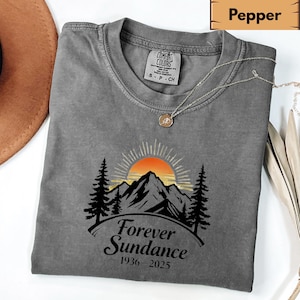 Könnte beinhalten: Ein graues Comfort Colors T-Shirt mit einem Berg- und Sonnenuntergang-Motiv. Die Grafik enthält den Text "Forever Sundance 1936-2025". Eine silberne Halskette mit Anhänger ist über dem Shirt drapiert.