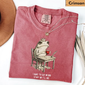 K&ouml;nnte beinhalten: Karmesinrotes T-Shirt mit einer Illustration einer gekr&ouml;nten Froschfigur, die an einem Schreibtisch sitzt, eine Tasse h&auml;lt und schreibt. Der Text "I HAVE TO SAY WEIRD STUFF OR I'LL DIE" ist darunter gedruckt. Eine silberne Halskette liegt auf dem Shirt.