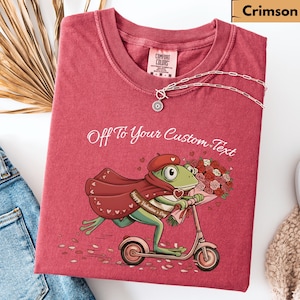 May include: Crimson t-shirt featuring a frog graphic on a scooter, with flowers, cape, and beret. The text "Off To Your Custom Text" is above the graphic. A silver necklace with a pendant is on the shirt.