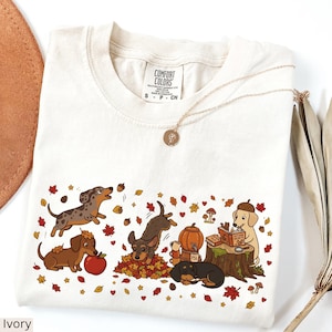 May include: An ivory t-shirt featuring a graphic print of various dachshunds in an autumn setting with falling leaves, acorns, a pumpkin, and a dog reading a book on a tree stump. The shirt has a Comfort Colors brand label at the neck.