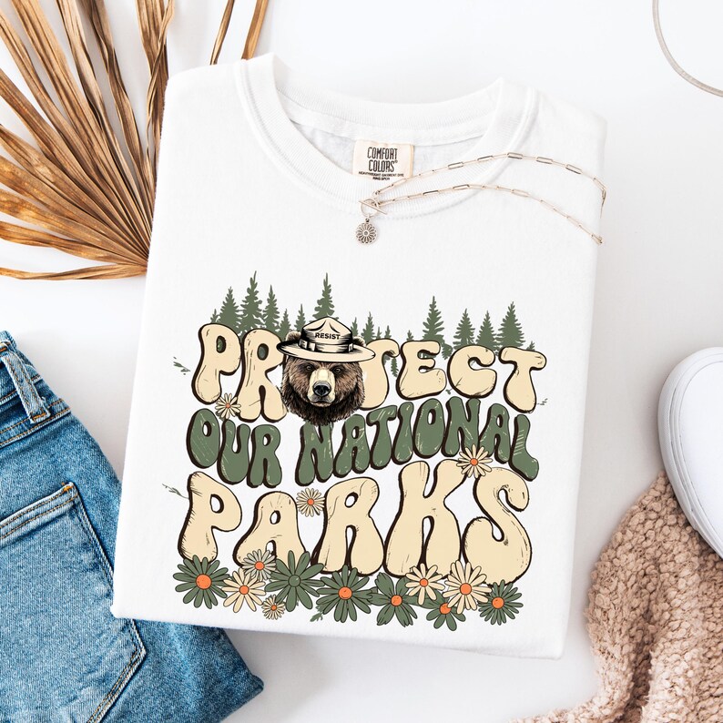 Puede incluir: Sudadera blanca Comfort Colors con el texto "PROTECT OUR NATIONAL PARKS" en una fuente retro, con un oso que lleva un sombrero de guardabosques. El dise&ntilde;o incluye &aacute;rboles y flores en tonos verdes y beige. Un collar plateado est&aacute; drapeado sobre la camisa.