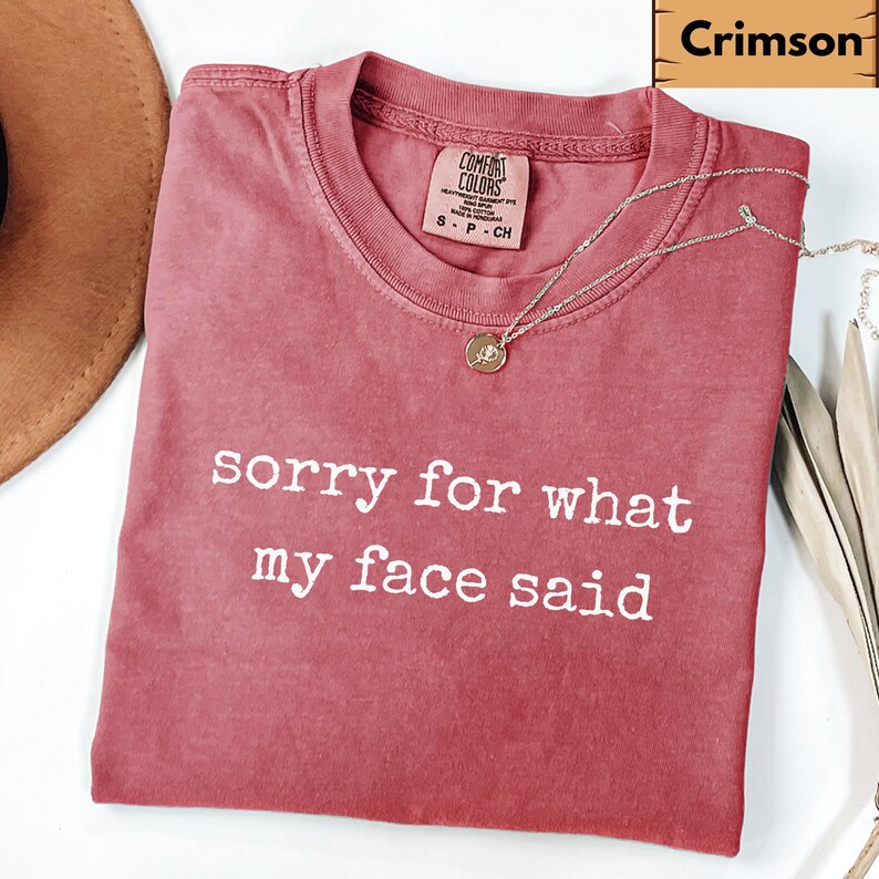May include: Crimson-colored t-shirt with the text "sorry for what my face said" in white. The shirt has a crew neck and a small tag on the upper left. A silver necklace with a round pendant is draped across the shirt.