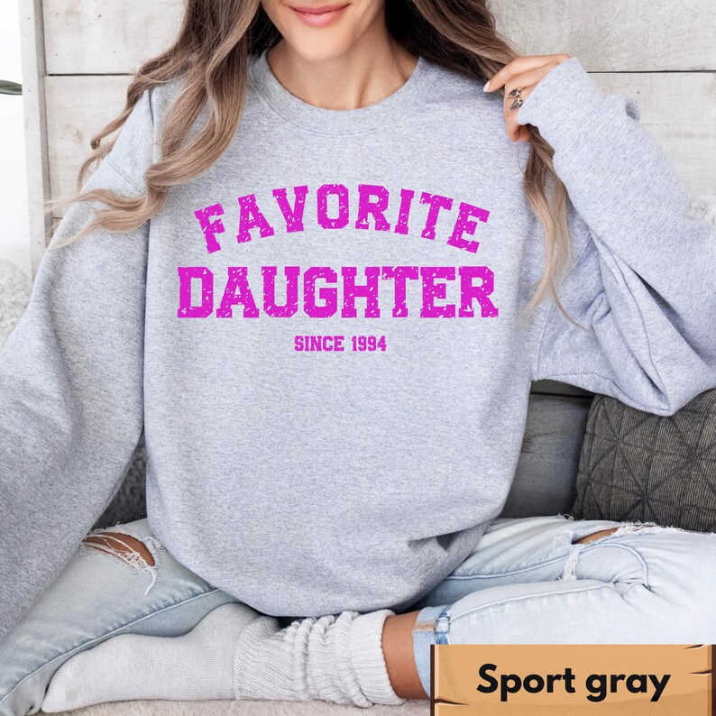 Puede incluir: Una sudadera gris claro con las palabras "FAVORITE DAUGHTER" en rosa brillante, arqueadas sobre el pecho, con "SINCE 1994" debajo. La sudadera es de cuello redondo.