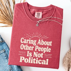 Puede incluir: Una camiseta roja Comfort Colors con la frase "Caring About Other People Is Not Political" en blanco. Una cadena plateada con un colgante floral descansa sobre la camiseta. Se ven unos vaqueros azules. La camiseta est&aacute; doblada.