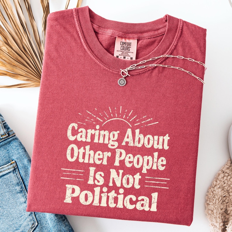 Puede incluir: Una camiseta roja Comfort Colors con la frase "Caring About Other People Is Not Political" en blanco. Una cadena plateada con un colgante floral descansa sobre la camiseta. Se ven unos vaqueros azules. La camiseta est&aacute; doblada.