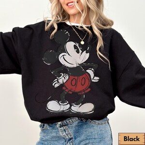 Puede incluir: Sudadera negra con un gr&aacute;fico desgastado de Mickey Mouse con pantalones cortos rojos. El personaje de dibujos animados est&aacute; de pie con las manos en las caderas. La sudadera tiene un cuello redondo blanco.