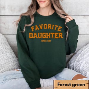Puede incluir: Sudadera verde bosque con el texto naranja "FAVORITE DAUGHTER" y "SINCE 1994" debajo. La sudadera es de color liso y tiene cuello redondo. La imagen muestra a una persona que lleva la sudadera.