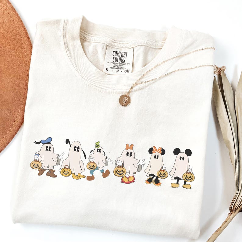 Op de afbeelding: Cr&egrave;mekleurig T-shirt met een ronde hals en korte mouwen. Het shirt heeft een rij cartoongeesten verkleed als populaire personages, elk met een pompoen. Een gouden ketting met een hanger is over het shirt gedrapeerd.