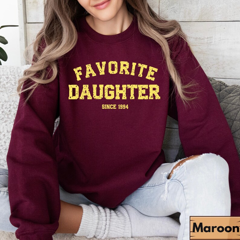 Puede incluir: Una sudadera granate con el texto "FAVORITE DAUGHTER" en amarillo, con "SINCE 1994" debajo. La sudadera es de color liso y tiene cuello redondo. La modelo lleva vaqueros de lavado claro.