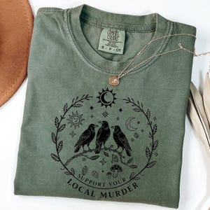 May include: An olive green t-shirt with a graphic of three black crows perched on a branch, surrounded by a wreath of celestial elements. The text "Support Your Local Murder" is below the graphic. A gold necklace and a brown hat are also visible.
