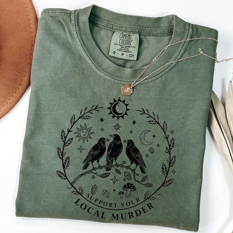 May include: An olive green t-shirt with a graphic of three black crows perched on a branch, surrounded by a wreath of celestial elements. The text "Support Your Local Murder" is below the graphic. A gold necklace and a brown hat are also visible.