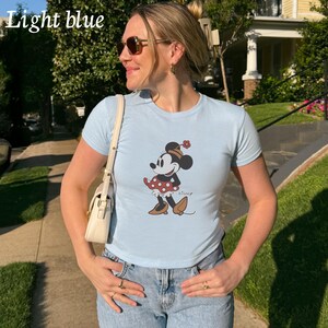 May include: Light blue t-shirt featuring a Minnie Mouse graphic. The cartoon character is wearing a red polka dot dress, a hat with a flower, and brown shoes. The shirt is light blue and has short sleeves. The word "Disney" is printed on the shirt.