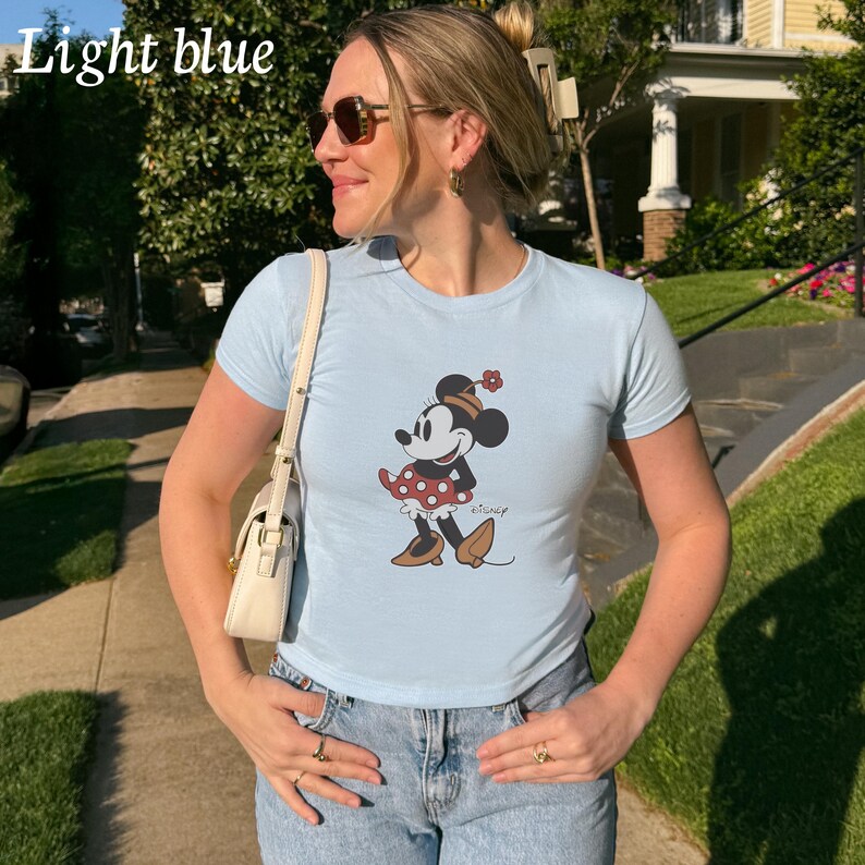 May include: Light blue t-shirt featuring a Minnie Mouse graphic. The cartoon character is wearing a red polka dot dress, a hat with a flower, and brown shoes. The shirt is light blue and has short sleeves. The word "Disney" is printed on the shirt.