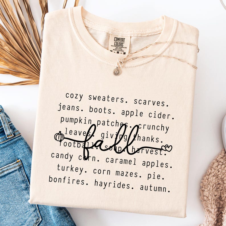 K&ouml;nnte beinhalten: Cremefarbenes T-Shirt mit dem Wort "fall" in Schreibschrift, umgeben von einer Liste von W&ouml;rtern zum Thema Herbst wie "cozy sweaters", "pumpkin patches" und "candy corn". Eine goldene Halskette mit Sonnenanh&auml;nger ist ebenfalls sichtbar.