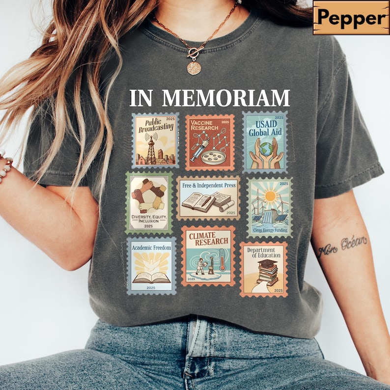 May include: Dark gray t-shirt featuring the words "IN MEMORIAM" above a collection of postage stamp-style graphics. The stamps depict themes like public broadcasting, vaccine research, and climate research. The shirt has a relaxed fit.