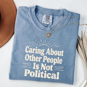 Puede incluir: Una camiseta azul con el texto "Caring About Other People Is Not Political" en una fuente retro. La camiseta tiene un dise&ntilde;o de sol sobre el texto. Un collar dorado con un colgante est&aacute; sobre la camiseta.