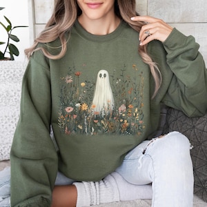 May include: Olive green sweatshirt featuring a whimsical ghost design. The ghost is depicted in a field of colorful wildflowers. The long-sleeved top is a comfortable, casual style, perfect for fall and Halloween.
