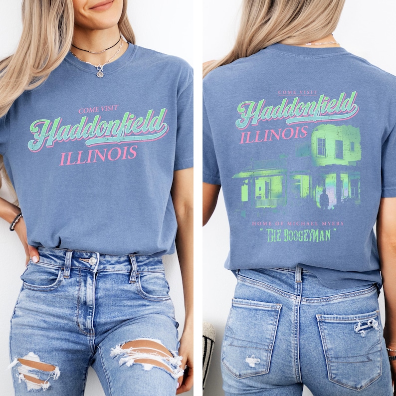 May include: Blue short-sleeved t-shirt with the text "Come Visit Haddonfield Illinois" in green and pink. The back of the shirt features a green and white graphic of a house and the text "Home of Michael Myers The Boogeyman."