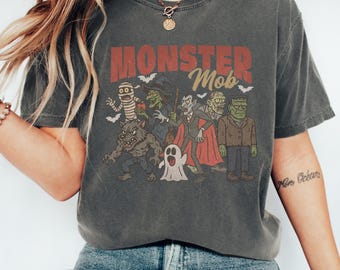May include: A charcoal gray short-sleeve t-shirt featuring the text "MONSTER Mob" in red and gold. The graphic displays a group of classic Halloween characters including a mummy, witch, vampire, werewolf, ghost, and Frankenstein's monster.