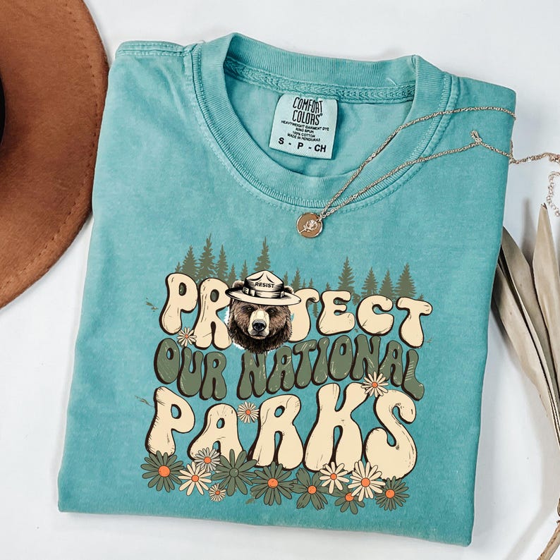 Puede incluir: Una camiseta turquesa Comfort Colors con el texto "PROTECT OUR NATIONAL PARKS" en una fuente retro, con un oso con sombrero de guardabosques. El dise&ntilde;o incluye &aacute;rboles y flores. Un collar dorado est&aacute; sobre la camiseta.