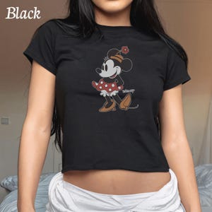 May include: Black crop top featuring a Minnie Mouse graphic. The cartoon character is depicted in a red polka dot skirt, brown shoes, and a matching flower on her hat. The word "Disney" is printed on the shirt.