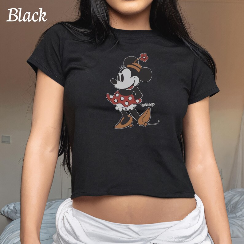 May include: Black crop top featuring a Minnie Mouse graphic. The cartoon character is depicted in a red polka dot skirt, brown shoes, and a matching flower on her hat. The word "Disney" is printed on the shirt.