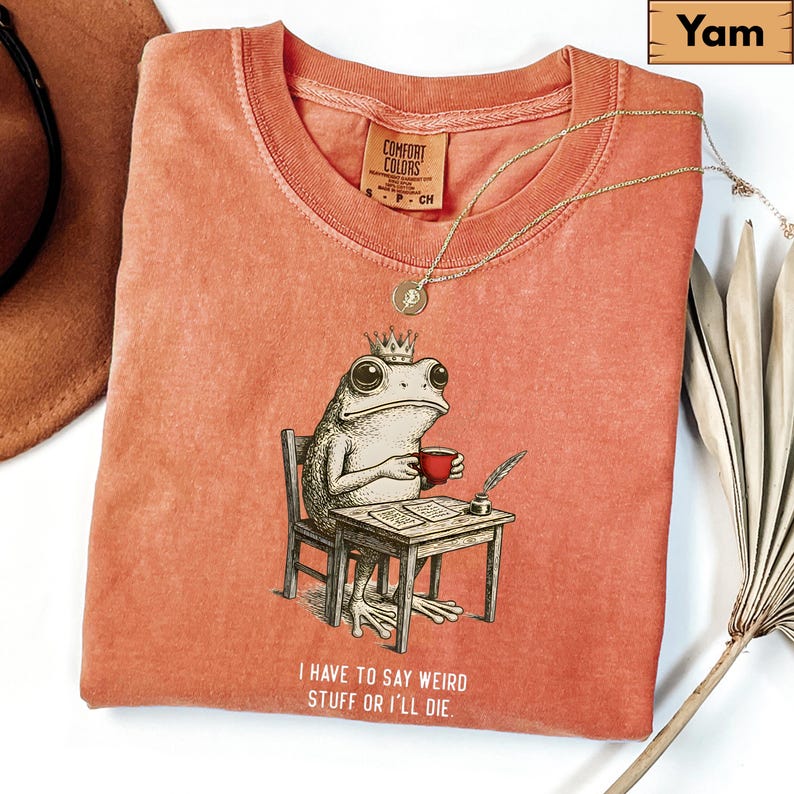 K&ouml;nnte beinhalten: Korallfarbenes T-Shirt mit einer skurrilen Illustration einer Froschk&ouml;nigin, die an einem Schreibtisch sitzt und eine rote Tasse h&auml;lt. Der Text "I HAVE TO SAY WEIRD STUFF OR I'LL DIE" ist darunter gedruckt. Eine goldene Halskette liegt auf dem Shirt.