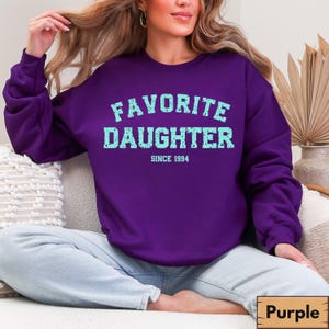 Puede incluir: Una sudadera morada con el texto "FAVORITE DAUGHTER" en azul claro, con "SINCE 1994" debajo. La sudadera es de color liso y tiene cuello redondo. La imagen tambi&eacute;n muestra unos vaqueros azules.