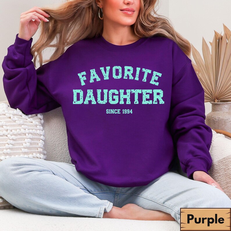 Puede incluir: Una sudadera morada con el texto "FAVORITE DAUGHTER" en azul claro, con "SINCE 1994" debajo. La sudadera es de color liso y tiene cuello redondo. La imagen tambi&eacute;n muestra unos vaqueros azules.