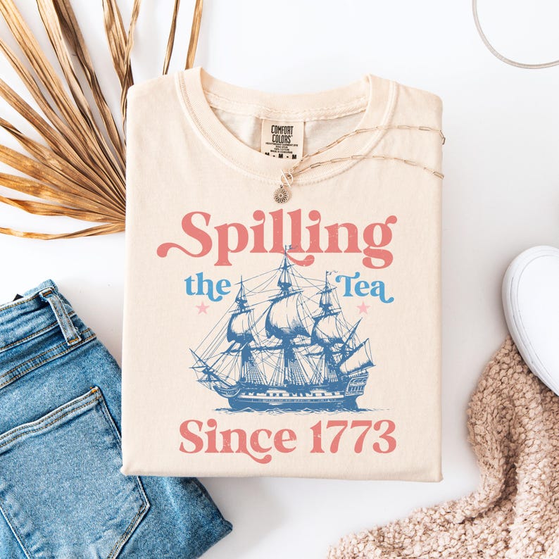May include: A beige t-shirt with a vintage-style graphic design. The design features a blue ship with the text "Spilling the Tea Since 1773" in pink and blue.