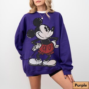 Puede incluir: Una sudadera morada con un gran gr&aacute;fico de Mickey Mouse en un estilo esbozado. Mickey est&aacute; en su atuendo cl&aacute;sico: pantalones cortos rojos, guantes negros y zapatos blancos. La sudadera tiene cuello redondo y mangas largas.