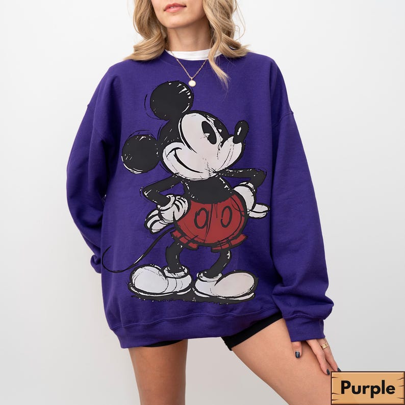 Puede incluir: Una sudadera morada con un gran gr&aacute;fico de Mickey Mouse en un estilo esbozado. Mickey est&aacute; en su atuendo cl&aacute;sico: pantalones cortos rojos, guantes negros y zapatos blancos. La sudadera tiene cuello redondo y mangas largas.