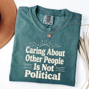 Puede incluir: Una camiseta verde azulado Comfort Colors con el texto "Caring About Other People Is Not Political" en blanco, con una fuente de estilo vintage. Un collar dorado con un colgante est&aacute; sobre la camiseta. Un sombrero marr&oacute;n es visible.