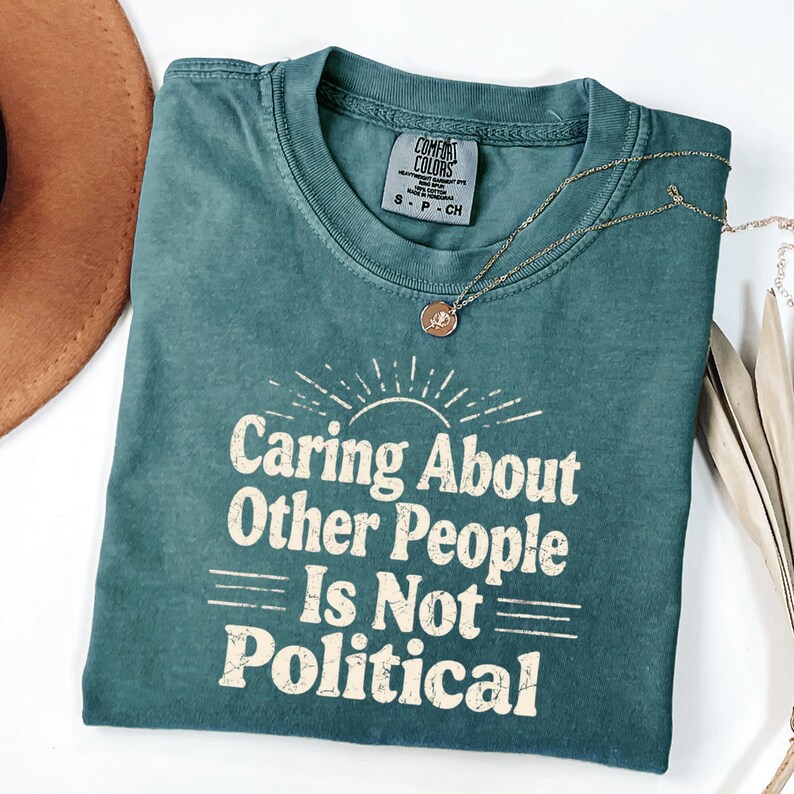 Puede incluir: Una camiseta verde azulado Comfort Colors con el texto "Caring About Other People Is Not Political" en blanco, con una fuente de estilo vintage. Un collar dorado con un colgante est&aacute; sobre la camiseta. Un sombrero marr&oacute;n es visible.