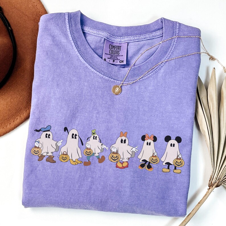 Op de afbeelding: Een lavendelkleurig Comfort Colors T-shirt met een rij cartoongeesten verkleed als populaire personages, elk met een pompoen. Het shirt heeft een ronde hals en korte mouwen. Een gouden ketting met een hanger ligt over het shirt.