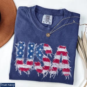 May include: A navy blue t-shirt with the word "USA" appliqued on the front in a distressed American flag design. The shirt has a crew neck and short sleeves. A gold necklace with a pendant is draped across the shirt.