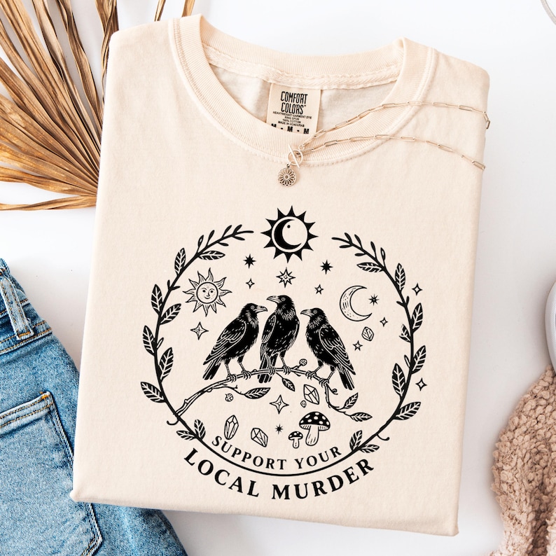 May include: Cream-colored Comfort Colors sweatshirt with a black graphic of three crows perched on a branch, surrounded by celestial elements. The text "Support Your Local Murder" is below the design. A gold necklace with a sun pendant is visible.