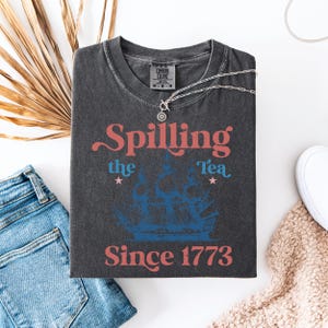 May include: A dark gray t-shirt with a vintage-style graphic that says "Spilling the Tea Since 1773". The graphic features a blue and white ship with red stars.