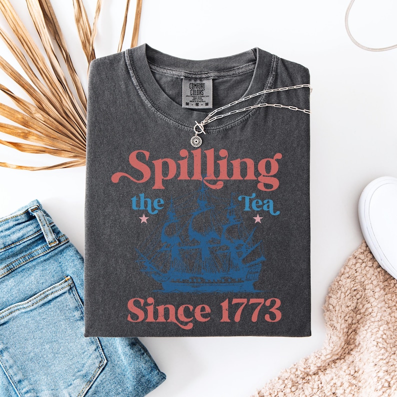 May include: A dark gray t-shirt with a vintage-style graphic that says "Spilling the Tea Since 1773". The graphic features a blue and white ship with red stars.