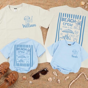 Könnte beinhalten: Zwei hellfarbene T-Shirts und zwei Baby-Bodys mit Strandmotiven. Die Erwachsenen-Shirts sind mit "William" und "Beach Crew" bedruckt. Die Baby-Bodys sind hellblau und mit "Noah" und "Beach Crew" bedruckt. Braune Sandalen und Sonnenbrillen sind ebenfalls zu sehen.