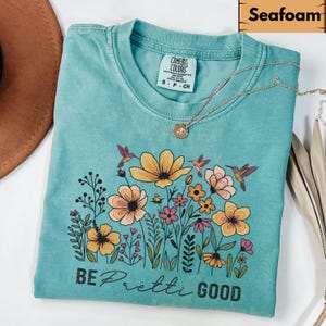May include: Seafoam green t-shirt with a floral design featuring yellow, pink, and peach flowers, and hummingbirds. The text "BE Pretti GOOD" is printed below the flowers. A gold necklace with a pendant is on the shirt.