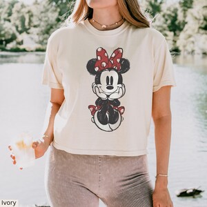 May include: Ivory-colored t-shirt featuring a distressed graphic of Minnie Mouse. Minnie is depicted in black and white with a red polka-dot bow. The shirt has a relaxed fit and short sleeves, perfect for casual wear.