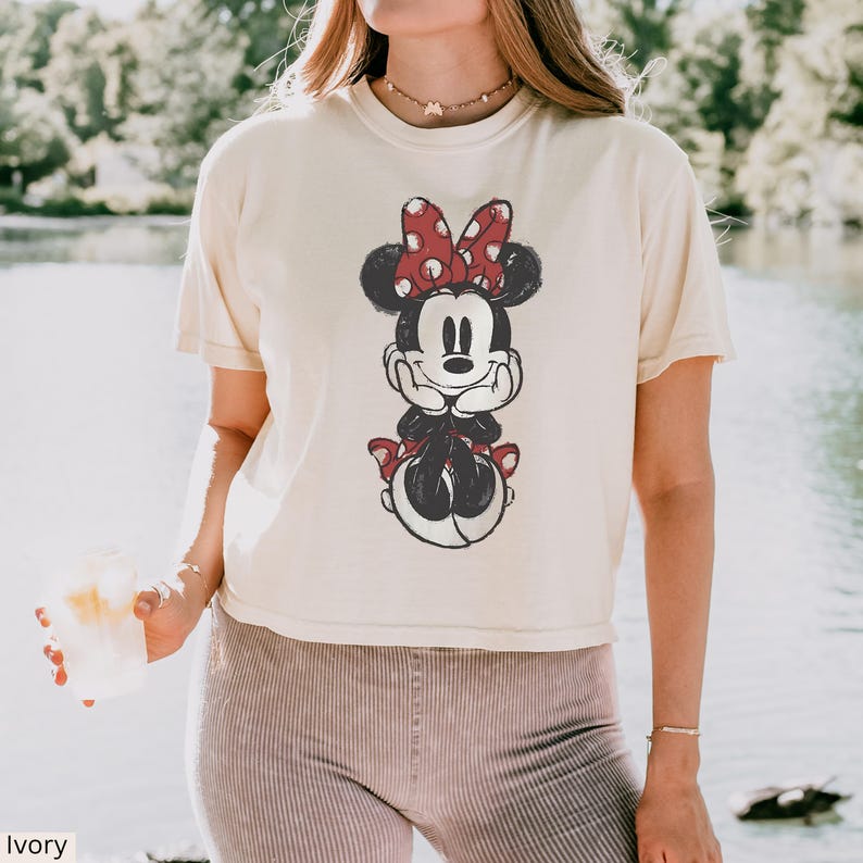 May include: Ivory-colored t-shirt featuring a distressed graphic of Minnie Mouse. Minnie is depicted in black and white with a red polka-dot bow. The shirt has a relaxed fit and short sleeves, perfect for casual wear.