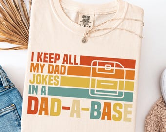 Könnte beinhalten: Cremefarbenes T-Shirt mit dem Text "I KEEP ALL MY DAD JOKES IN A DAD-A-BASE" in Retro-Design. Die Grafik enthält ein Disketten-Symbol und horizontale Streifen in Orange, Gelb und Türkis.