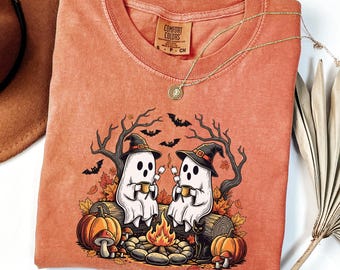 Può includere: T-shirt Comfort Colors arancione con grafica a tema Halloween. La grafica presenta due fantasmi dei cartoni animati con cappelli da strega, che tengono tazze e seduti vicino a un falò. Sono incluse anche zucche, pipistrelli e un gatto nero. Una collana d'oro è drappeggiata sulla maglietta.
