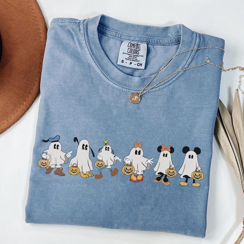 Op de afbeelding: Een lichtblauw Comfort Colors T-shirt met een ronde hals en korte mouwen. Het heeft een rij cartoongeesten verkleed als Disney-personages, elk met een pompoen. Een gouden ketting is over het T-shirt gedrapeerd.