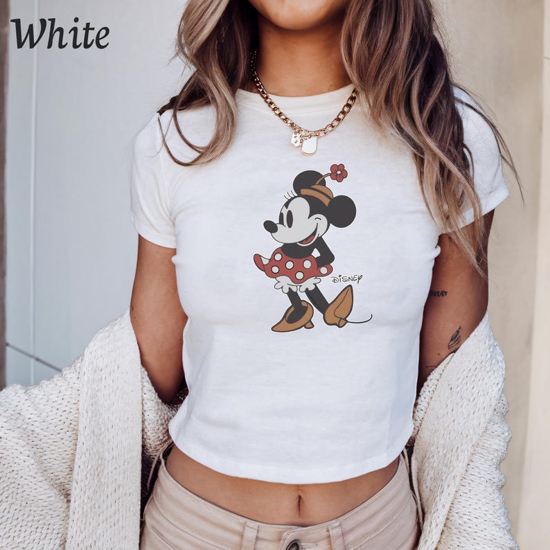 May include: White crop top featuring a Minnie Mouse graphic. The cartoon character is depicted in a red polka dot skirt and brown shoes. The word "Disney" is printed below the character.