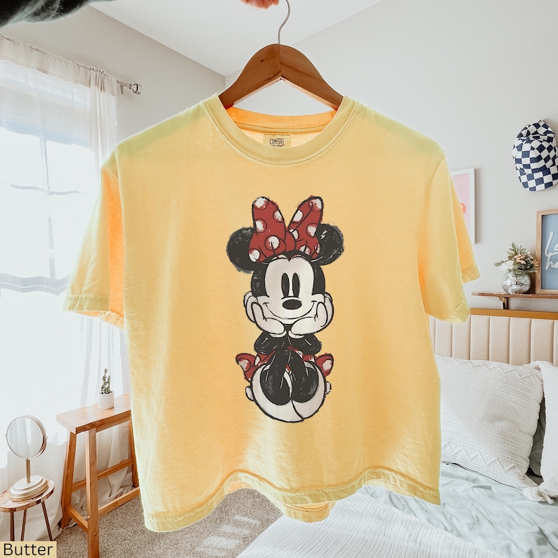 May include: Butter yellow t-shirt featuring a graphic of Minnie Mouse. The cartoon character is depicted with a red polka dot bow, black hair, and a smiling face. The shirt is hanging on a wooden hanger.